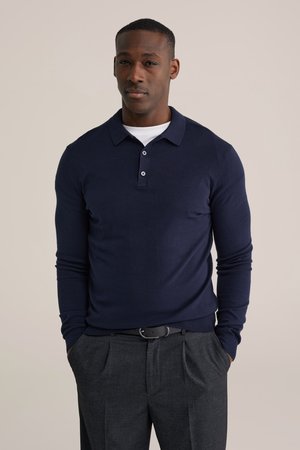 Maglione polo a maniche lunghe blu navy con due bottoni al colletto, abbinato a pantaloni grigi plissettati e cintura nera. Tessuto liscio.