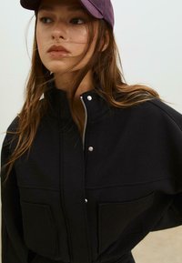 Črna zip-up jakna z dvema sprednjima žepoma, visokim ovratnikom in pritisnimi gumbki; nosi se z temno vijolično kapo; mehak material, sproščen kroj.