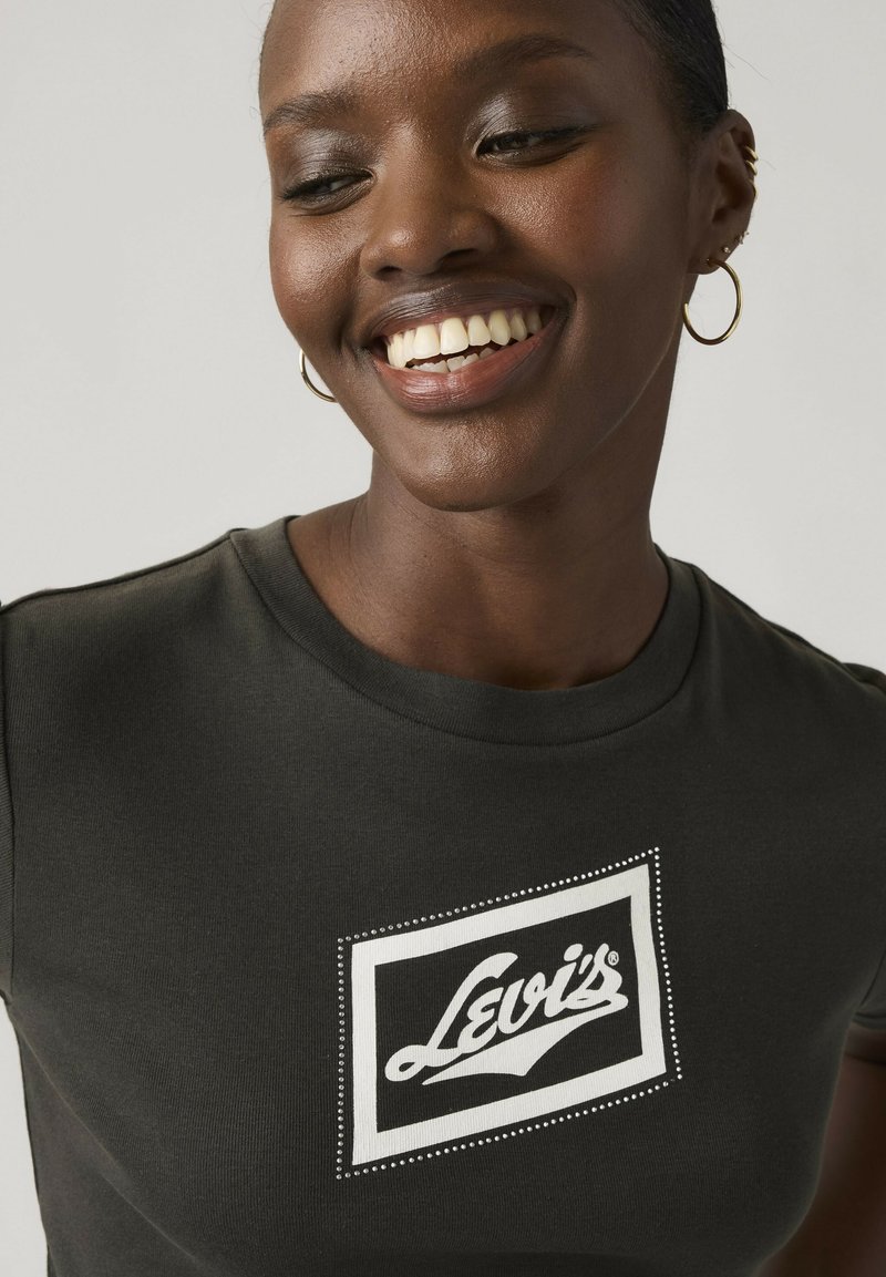 Glunderende vrouw met een donker Levi's T-shirt en gouden oorringen, naar beneden kijkend tegen een effen lichte achtergrond.