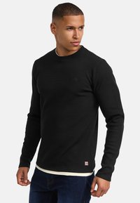 Camisa negra de manga larga con textura acanalada y cuello redondo, con un pequeño logo en el pecho. Lleva puesta sobre una camisa de color claro y unos vaqueros azules.