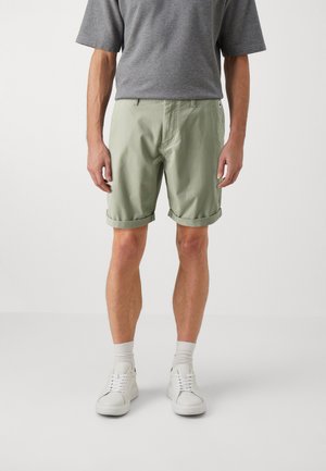 Pantalones cortos de algodón verde claro con dobladillos enrollados, un frente plano, bolsillos laterales y una pequeña etiqueta de logo, combinados con zapatillas blancas y una camiseta gris.