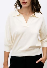 Pull en tricot de couleur crème avec un col en V et des manches courtes bouffantes, fabriqué en tissu doux, avec des poignets côtelés et une coupe décontractée.