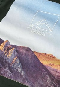 Koszulka bawełniana z grafiką gór na tle w gradientowych odcieniach niebieskiego. Wzór zawiera białe elementy geometryczne oraz tekst.