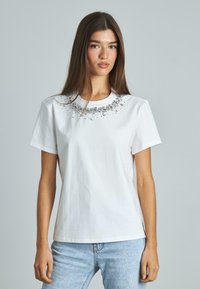 Camiseta blanca de manga corta hecha de algodón, con un escote adornado con embellishments plateados y transparentes, textura suave y ajuste clásico.