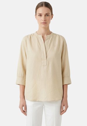 Donna che indossa una camicetta beige a maniche lunghe in lino con scollo a fessura e pantaloni bianchi su uno sfondo chiaro e semplice.