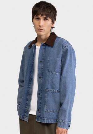 Νέος άνδρας με κοντά καστανά μαλλιά, φοράει μπλε τζιν μπουφάν με καφέ γιακά, λευκό πουκάμισο και καφέ παντελόνι, μπροστά σε ένα απλό φόντο.