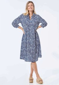 Kleid aus blauem und weißem gemustertem Stoff mit Knopfleiste, langen Ärmeln und tailliertem Schnitt. Kombiniert mit hellen, gewebten Plateau-Sandalen.