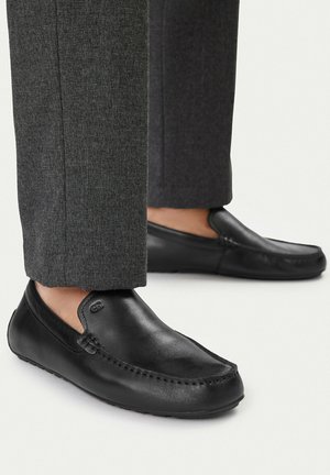 Czarne wsuwane mokasyny ze skóry noszone z szarymi teksturowanymi spodniami, pokazane z boku na jasnym tle.