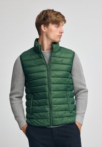 Grüne Steppweste mit Reißverschluss, gesteppter Optik und hohem Kragen. Über einem grauen strukturierten Pullover getragen, der ein einfaches besticktes Logo trägt.