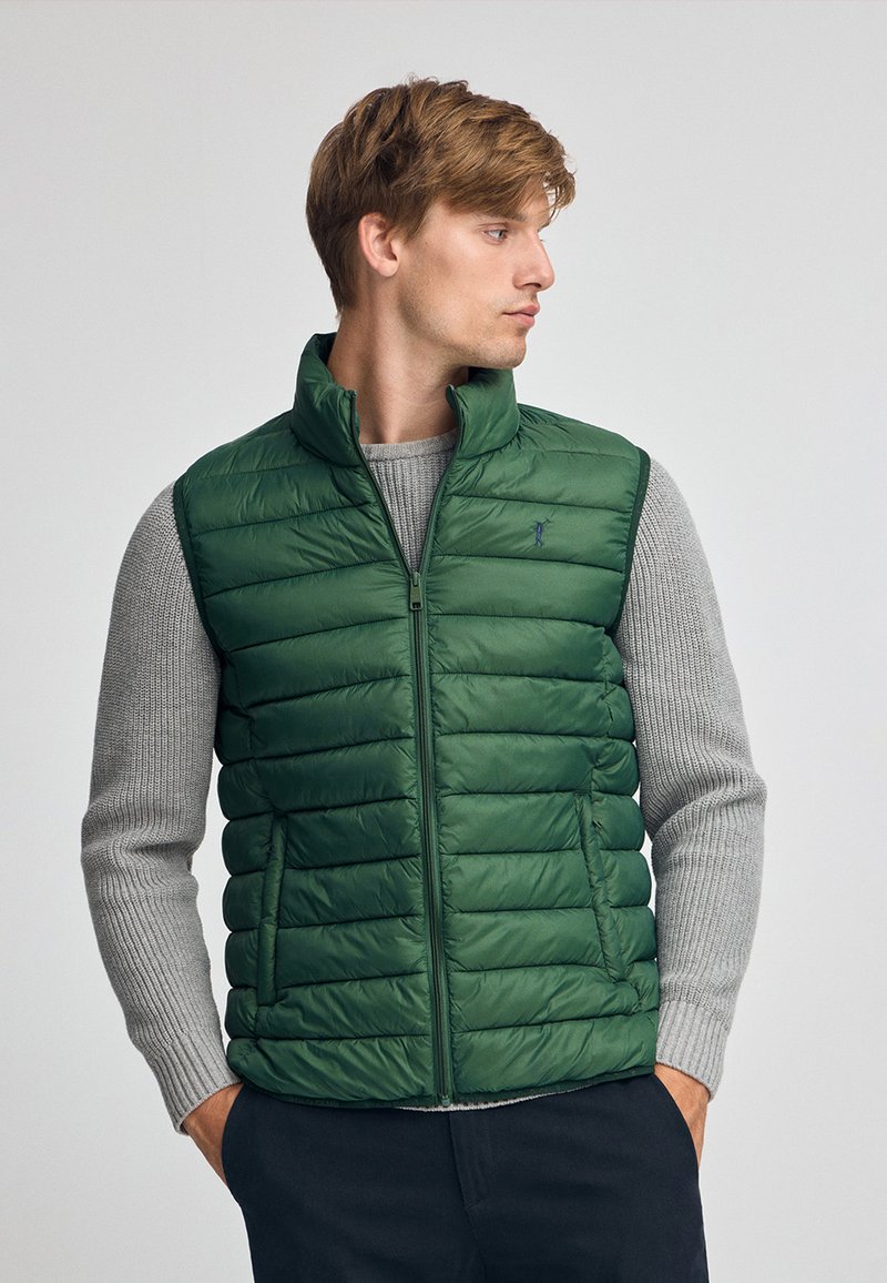 Grüne Steppweste mit Reißverschluss, gesteppter Optik und hohem Kragen. Über einem grauen strukturierten Pullover getragen, der ein einfaches besticktes Logo trägt.