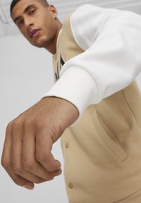 Felpa in jersey beige con maniche in tessuto bianco. Presenta polsini a coste, chiusura frontale con bottoni e tasche laterali. Mano sollevata in posizione di pugno.
