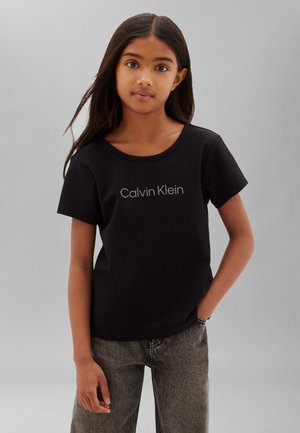 Mladé dievča s dlhými tmavými vlasmi, ktoré nosí čierne tričko Calvin Klein a sivé džínsy, stojí pred jednoliatym sivým pozadím.