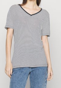 Žena oblečená v černém a bílém vodorovně pruhovaném tričku s výstřihem do V, v kombinaci s modrými džíny, má ruce uvolněně podél těla.