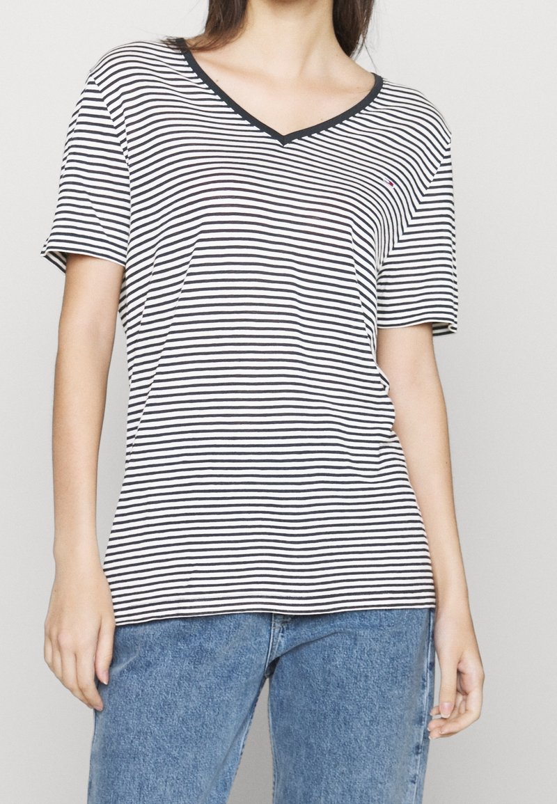 Žena oblečená v černém a bílém vodorovně pruhovaném tričku s výstřihem do V, v kombinaci s modrými džíny, má ruce uvolněně podél těla.