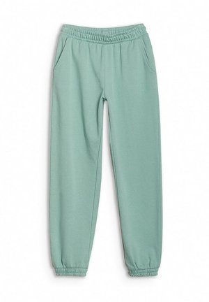 Jeune personne debout portant un sweat à capuche vert clair et un pantalon de survêtement assorti avec des baskets grises sur fond uni.