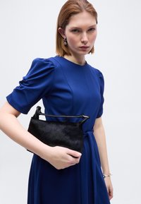 Γυναίκα που φοράει μπλε φόρεμα με φουσκωτά κοντά μανίκια κρατά μια μικρή μαύρη τσάντα χειρός με λαβή πάνω, πάνω σε απλό ανοιχτό φόντο.