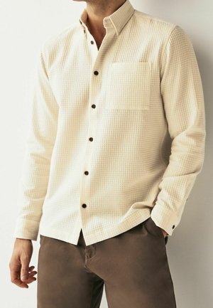 Light cream long-sleeve shirt with a textured woven pattern, button-up front, and single chest pocket. Dark buttons contrast with the fabric.