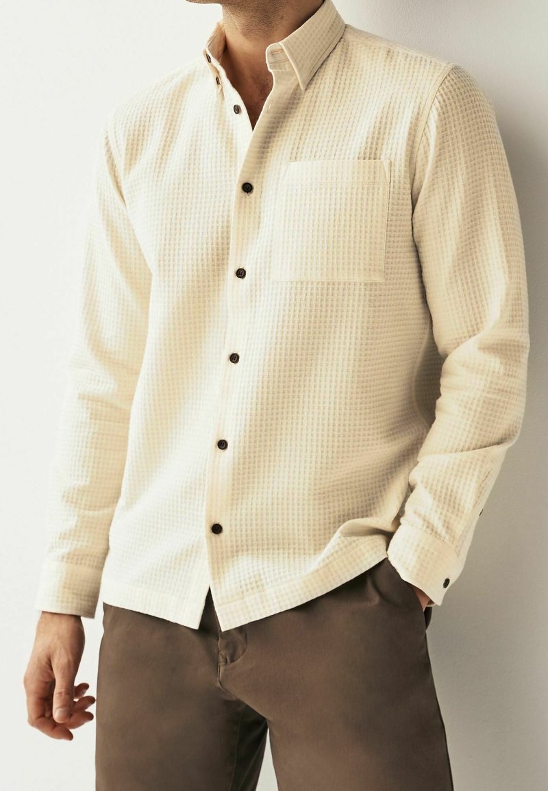 Chemise à manches longues couleur crème clair avec un motif tissé texturé, fermeture à boutons sur le devant et poche poitrine unique. Des boutons foncés contrastent avec le tissu.