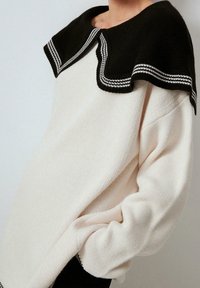 Gestrickter Pullover mit einem cremefarbenen Körper und einem schwarzen Kragen mit weißem Rand. Weiche Textur, übergroße Passform und abgefallene Schultern.