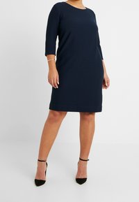 Femme portant une robe bleu marine arrivant au genou avec des manches trois-quarts et des escarpins noirs à bride cheville, se tenant devant un fond blanc uni.