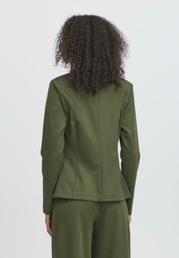 Giacca su misura verde oliva con maniche lunghe, design strutturato e dettagli delle cuciture sul retro. Realizzata in tessuto morbido e testurizzato.