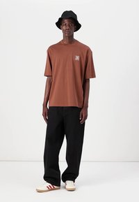 Καφέ βαμβακερό μπλουζάκι με κοντά μανίκια και μικρό λογότυπο. Φορεμένο με μαύρο παντελόνι σε φαρδιά γραμμή και μαύρο καπέλο τύπου bucket. Λευκά παπούτσια με κόκκινες ρίγες.