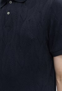 Πολό μπλε ναυτικού με υφασμένο μοτίβο φύλλων, δύο λευκά κουμπιά, κοντά μανίκια και διακριτικό κεντητό λογότυπο Guess στο στήθος.