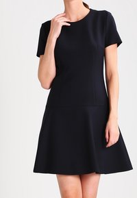Černé šaty s krátkým rukávem, těsně přiléhající k tělu a s rozšířenou sukní. Materiál vypadá hladce, má kulatý výstřih a žádné viditelné vzory či ozdoby.