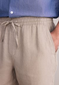 Pantaloni in lino beige chiaro con vita elasticizzata, coulisse e tasche laterali. Il tessuto presenta un aspetto liscio e leggermente strutturato.