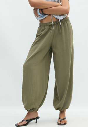 Femme portant un pantalon ample vert olive avec cordon de serrage et poignets élastiques, ainsi que des sandales à talons noires, les bras croisés sur le ventre.