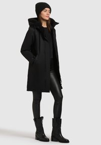 Zwarte parka met een met bont gevoerde capuchon, gecombineerd met een zwarte top, strakke leren broek en kniehoge zwarte laarzen. Heeft een gladde, glanzende afwerking.