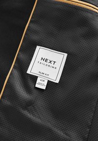 Czarna tkanina o siateczkowej strukturze, z białą metką z napisem "NEXT TAILORING", rozmiar "40R" oraz "SLIM FIT". Akcentowana złotą lamówką.