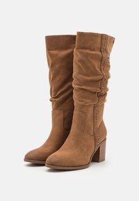 Bottes montantes en daim marron avec un design décontracté, des accents cousus et un talon carré. Texture lisse et bout arrondi.