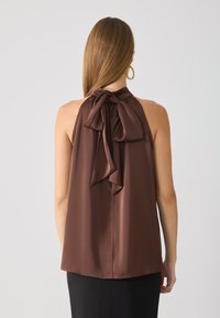 Bruine halter-top met een grote, geknoopte strik bij de hals en een gladde textuur, gecombineerd met een zwarte rok, gezien vanuit de rug.