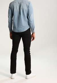 Ljust blå denimskjorta med långa ärmar och knappmuddar, ihopparad med svarta slim-fit jeans och vita sneakers, sedd bakifrån.