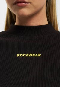 Svart ribbestrikket høyhalset genser med en gul brodert "ROCAWEAR"-logo på brystet. Nærbilde viser tekstur og detalj av halslinjen.