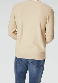 Pull en tricot beige avec une finition texturée, à manches longues et col rond, associé à un jean en denim bleu avec des détails sur les poches arrière.