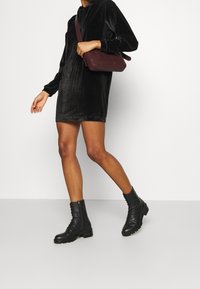 Abito nero in velluto con cappuccio e maniche lunghe, abbinato a una borsa a tracolla color borgogna e stivali neri con lacci alla caviglia. Tessuto morbido, lunghezza sopra il ginocchio.