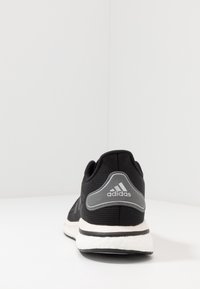 Chaussure de course Adidas noire vue du talon, avec une semelle blanche amortissante et un logo Adidas réfléchissant à l'arrière.