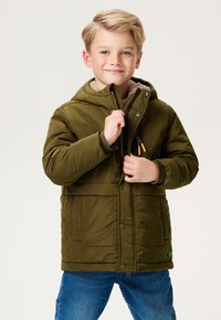 Blond chłopiec uśmiechający się, noszący oliwkowozieloną puchową kurtkę z kapturem i niebieskie dżinsy, zapinający suwak kurtki na jednolitym tle.