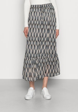 Femme portant une jupe midi à motif noir et blanc avec un ourlet à volants, un haut à manches longues blanc et des baskets blanches, debout sur un sol gris.