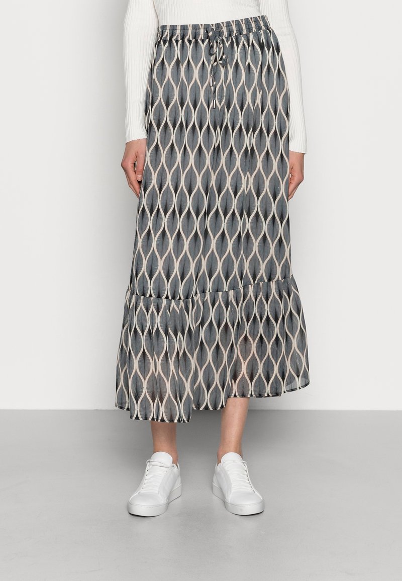 Femme portant une jupe midi à motif noir et blanc avec un ourlet à volants, un haut à manches longues blanc et des baskets blanches, debout sur un sol gris.