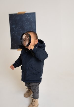 Un tout-petit portant un manteau bleu marine et des bottes tient une loupe devant son visage, debout près d'une affiche sombre étoilée fixée au mur avec du ruban adhésif.