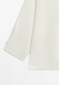 Kremowy sweter z długim rękawem, zbliżenie na lewy rękaw i boczny szew pokazujący prosty ścieg.