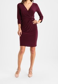 Viininpunainen mekko V-kaula-aukkolla, 3/4-hihasilla, sivuiltaan laskostettu ja pyöreä korostus. Pehmeää, vartalonmyötäistä kangasta polvenpituudessa.