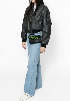 Giacca bomber in pelle nera, jeans a zampa d'elefante blu e una piccola borsa a tracolla nera con un accento a stampa leopardata verde. Scarpe bianche.