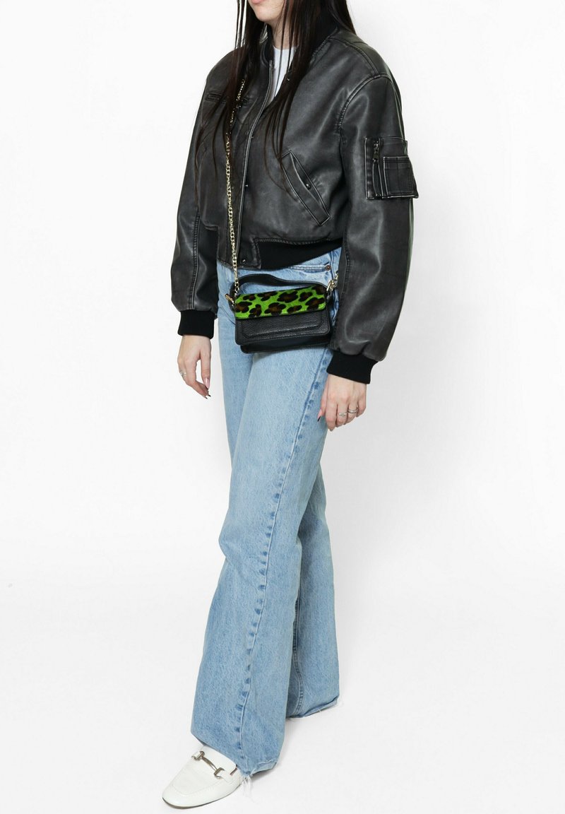 Zwarte leren bomberjack, blauwe flared jeans en een kleine zwarte crossbody tas met een groene luipaardprintaccent. Witte schoenen.