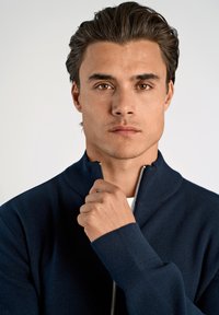 Laivastonsininen vetoketjullinen villapaita, jossa on korkea kaulus, ribbetyt hihansuut ja sileä rakenne. Mallin käsi pitää vetoketjusta kiinni, mikä korostaa design-yksityiskohtaa.