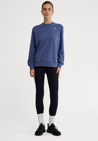 Felpa blu con polsini a coste, abbinata a leggings neri e calzini bianchi. La felpa presenta un piccolo logo sul petto.