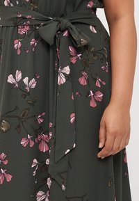 Green floral dress with pink flowers, featuring a belted waist and flowing fabric. The design includes detailed floral patterns and soft texture.